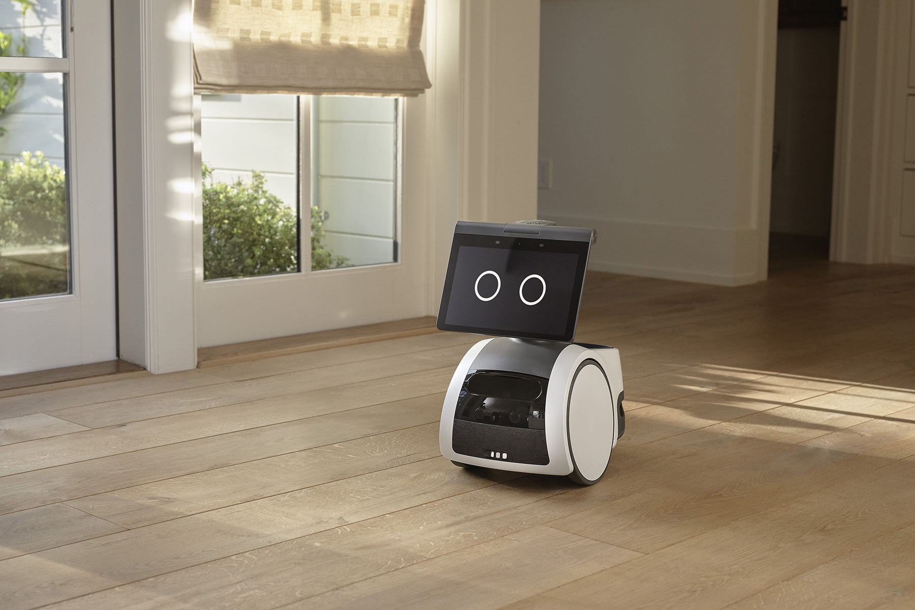 Amazon Astro home robot, white and gray rolling body with screen face, on a sunlit wooden floor